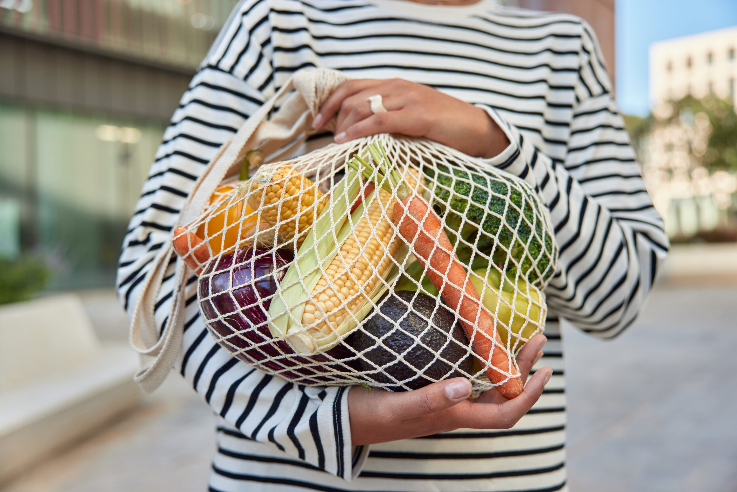 Cropped shot of faceless young woman walks with purchases carries fresh vegetables in eco friendly string mesh bag dressed in striped jumper poses against street background. Zero waste concept